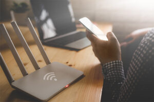 An image featuring a person using his phone with his wifi router and laptop in the background